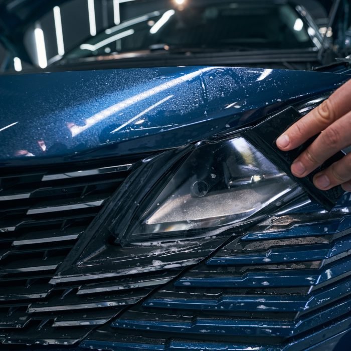 Closeup car service worker installing PPF with squeegee on headlight Closeup car service worker installing PPF with squeegee on headlight